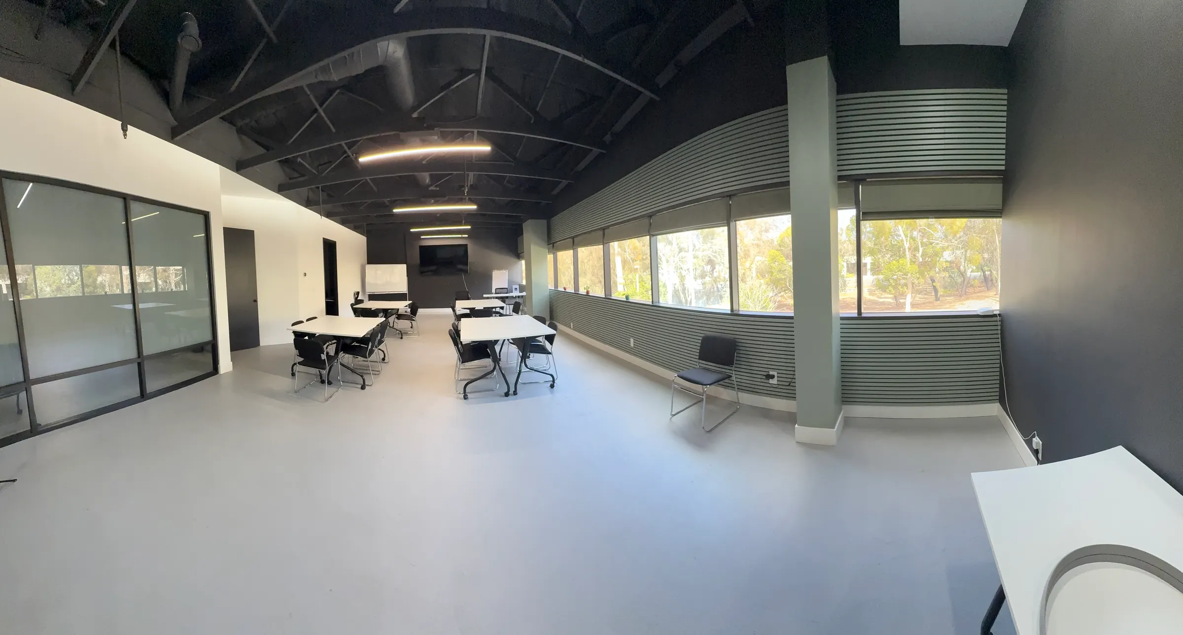 The training room edge to edge — black open ceiling, pale floor, a few white tables in the middle, and a wall of windows onto eucalyptus trees.