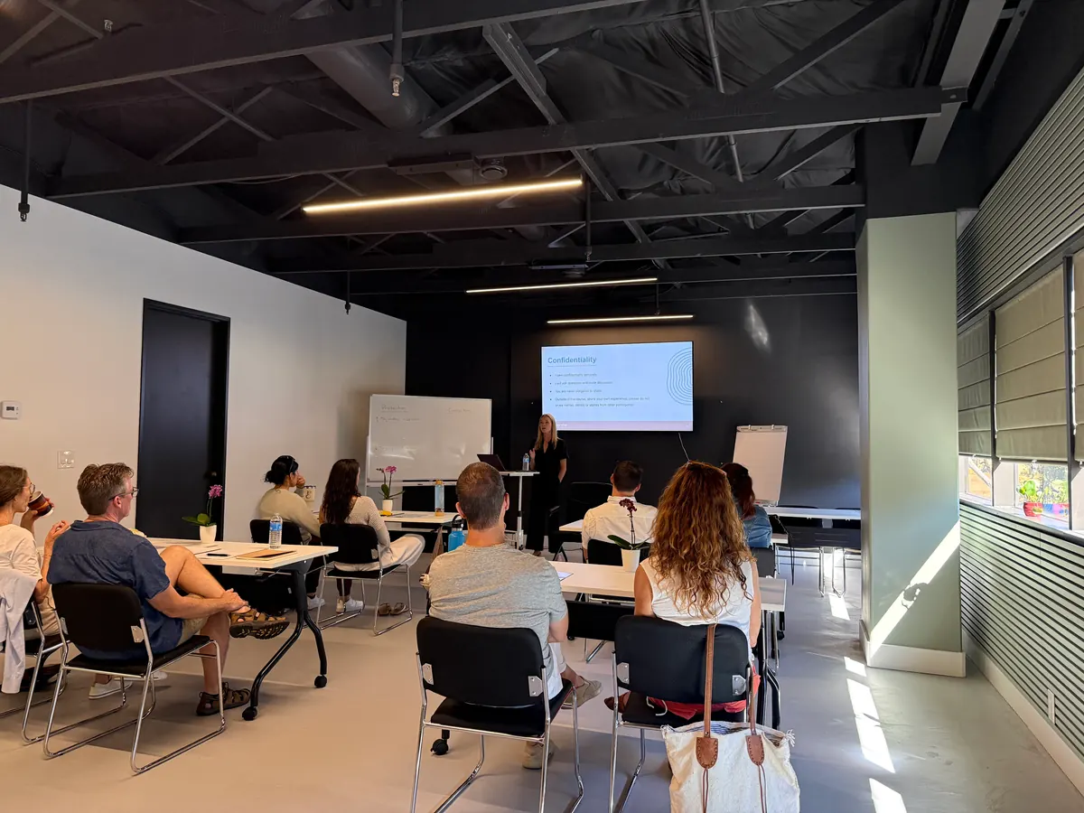A speaker presenting slides on the wall-mounted screen to an audience seated at white tables.